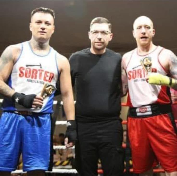 Two boxers in SORTED sponsored vests with the referee after a bout